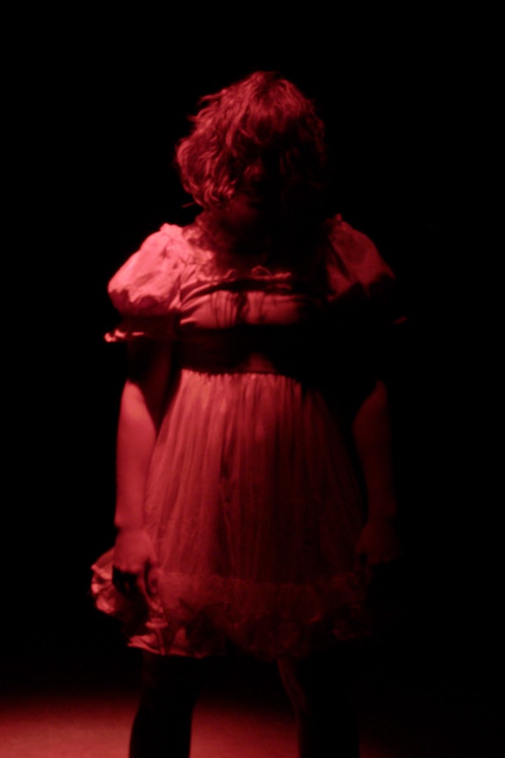 Photo from a Music Video, A Girl in a short puffy dress stands in a harsh shadow and an ominous red light.